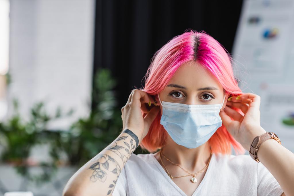 tetovaný mladý manažer s růžovými vlasy nosí lékařskou masku v kanceláři - Fotografie, Obrázek
