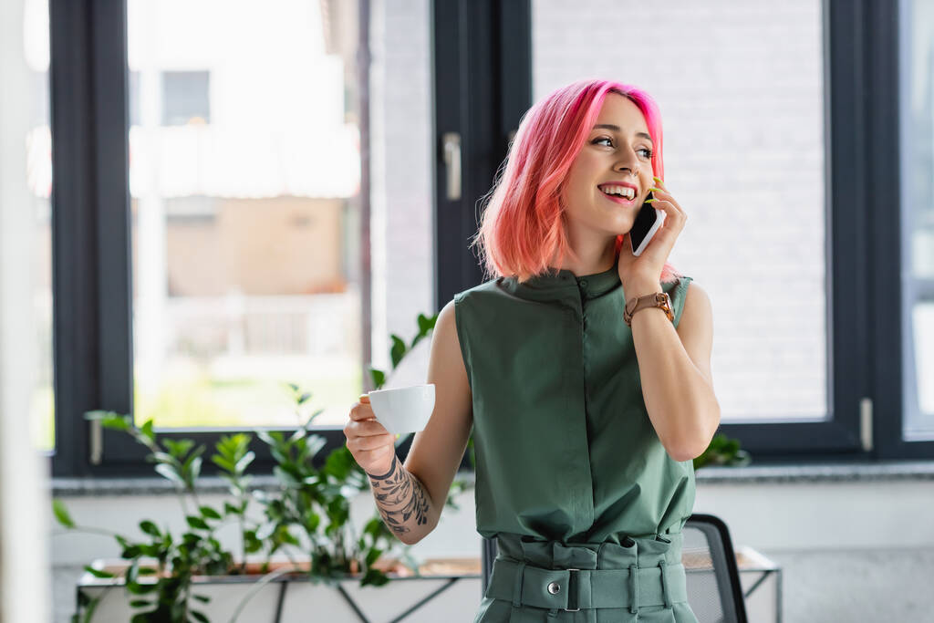 cheerful businesswoman with pink hair and piercing holding cup of coffee while talking on smartphone in office - Photo, Image