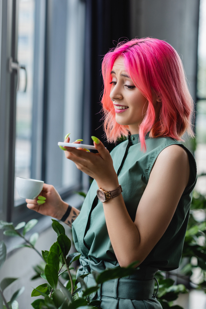 mulher de negócios alegre com cabelo rosa e piercing segurando xícara de café durante a gravação de mensagem de voz no smartphone - Foto, Imagem