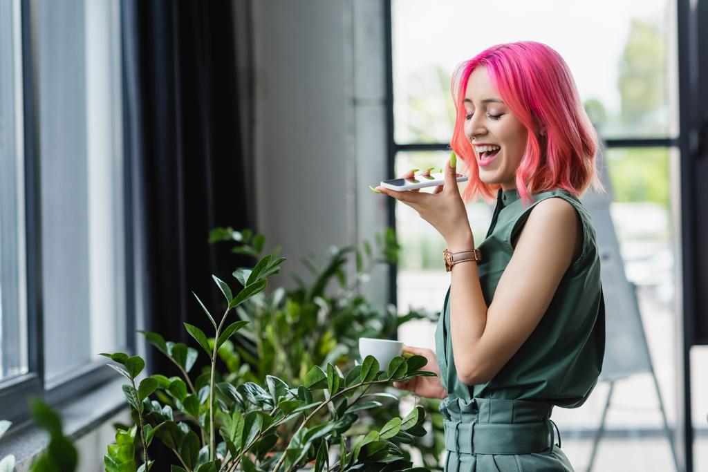 позитивна бізнес-леді з рожевим волоссям і пірсингом тримає чашку кави під час запису голосового повідомлення на смартфон
 - Фото, зображення