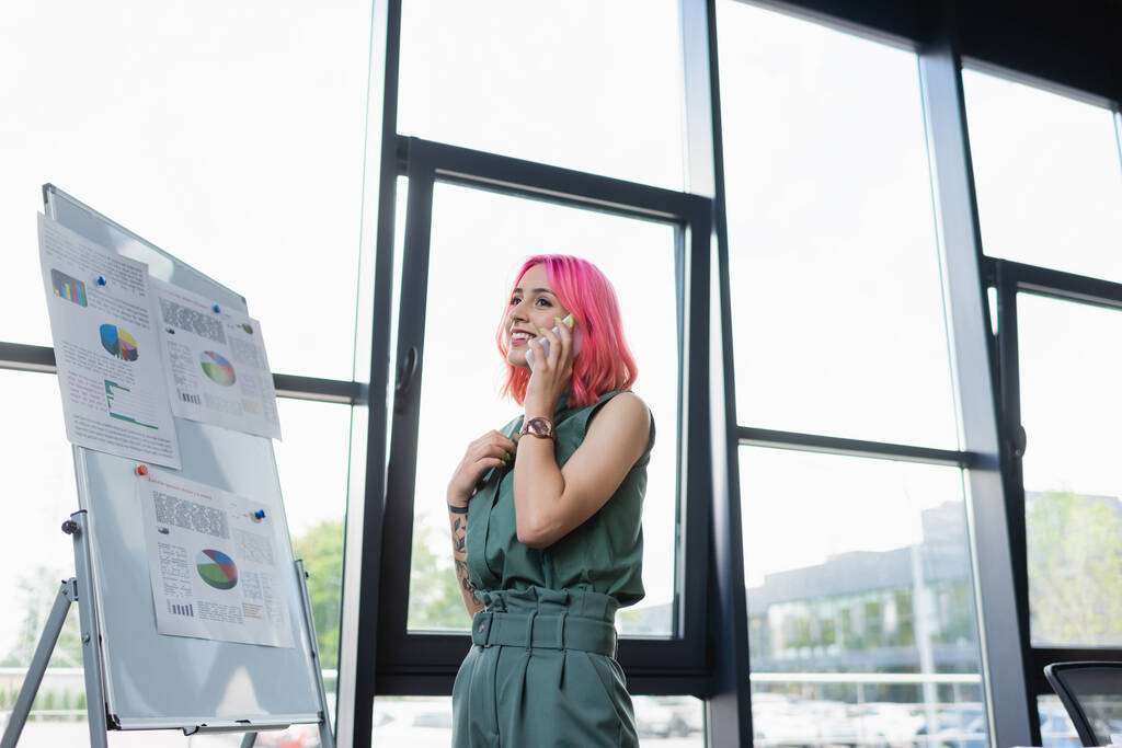 Ευτυχισμένη επιχειρηματίας με ροζ μαλλιά μιλάει στο κινητό κοντά στο διάγραμμα flip με διαγράμματα  - Φωτογραφία, εικόνα