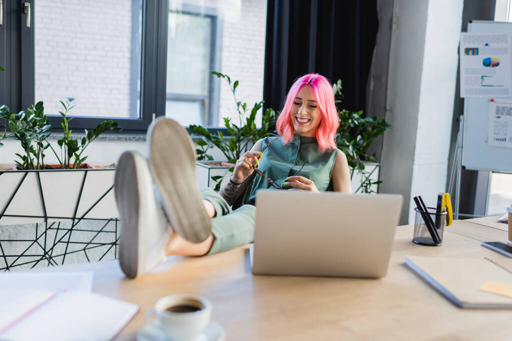 счастливая деловая женщина с розовыми волосами, держащая очки, глядя на ноутбук  - Фото, изображение