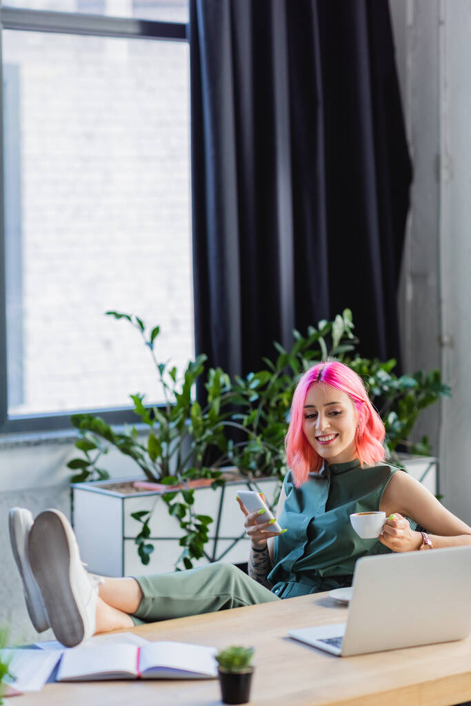 szczęśliwy businesswoman z różowymi włosami trzymając smartfona i filiżankę kawy patrząc na laptopa  - Zdjęcie, obraz