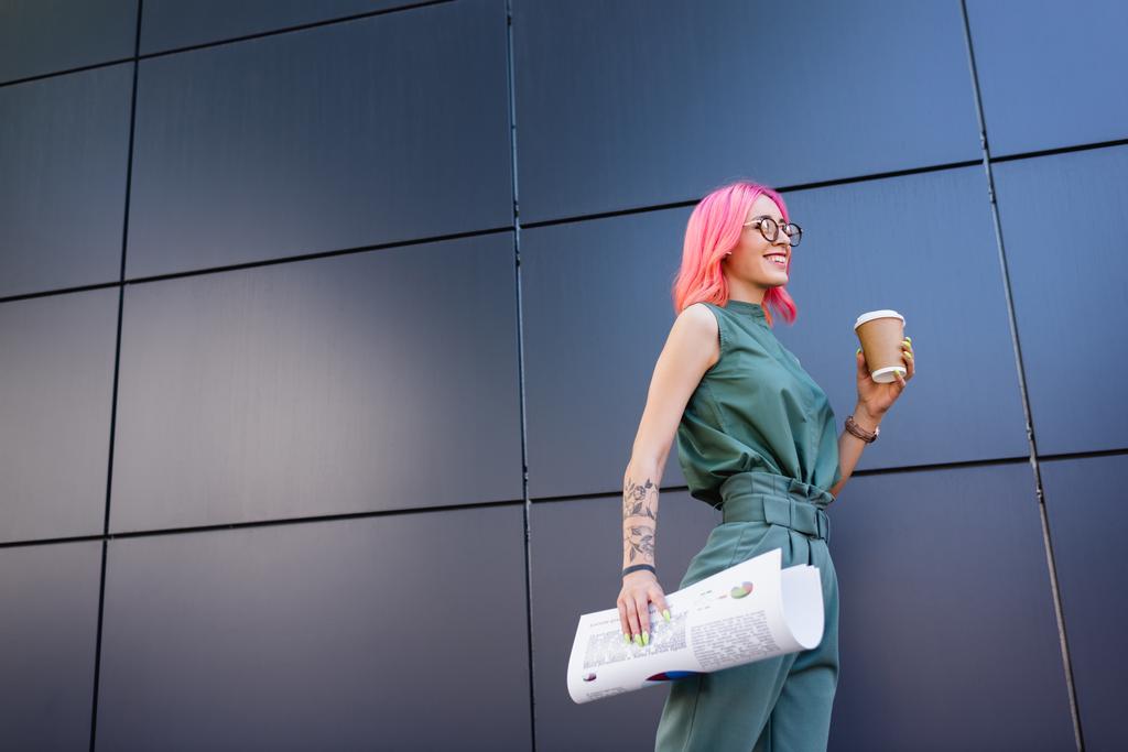 donna d'affari felice e tatuata con i capelli rosa che tengono tazza di carta e grafici all'esterno  - Foto, immagini