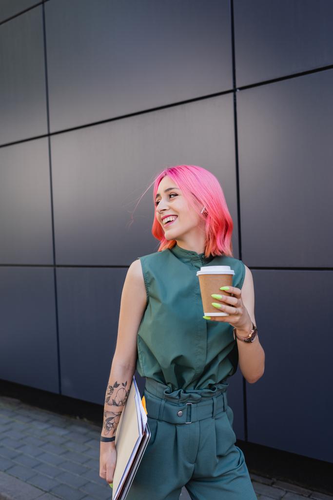 mosolygós üzletasszony rózsaszín hajjal és fülhallgatóval kezében kávé menni és mappák - Fotó, kép