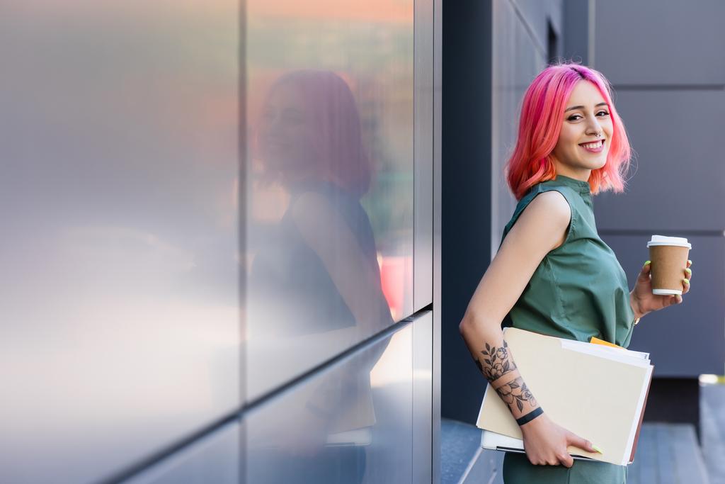 tattooed and happy businesswoman with pink hair holding folder, laptop and coffee to go outside - Photo, Image