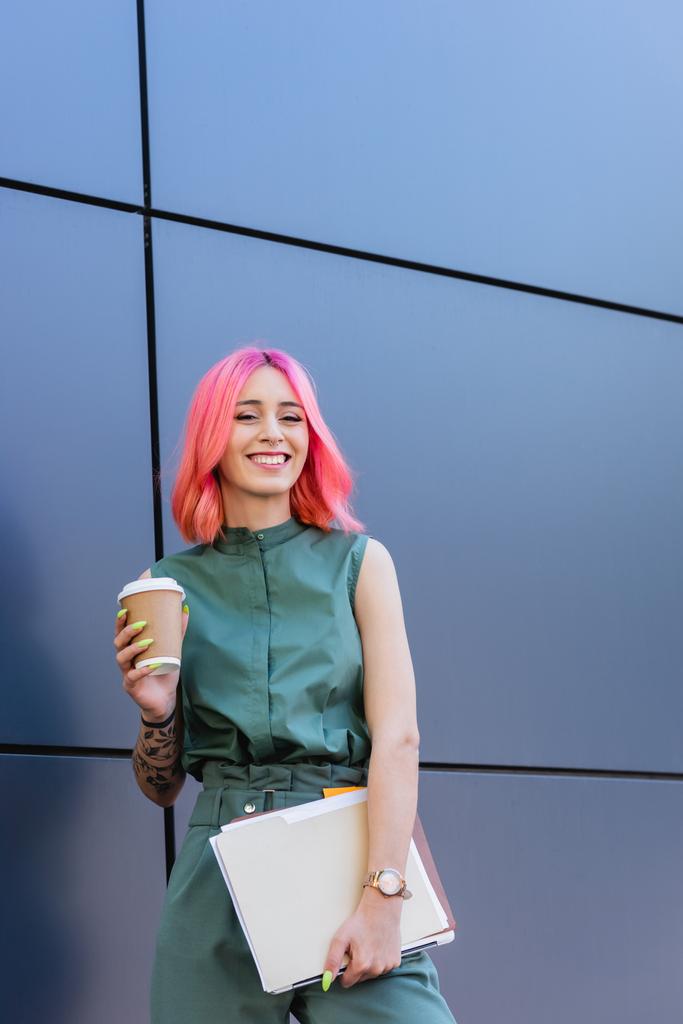 wytatuowana i pozytywna bizneswoman z różowymi włosami trzymająca folder, laptop i kawę, aby wyjść na zewnątrz - Zdjęcie, obraz