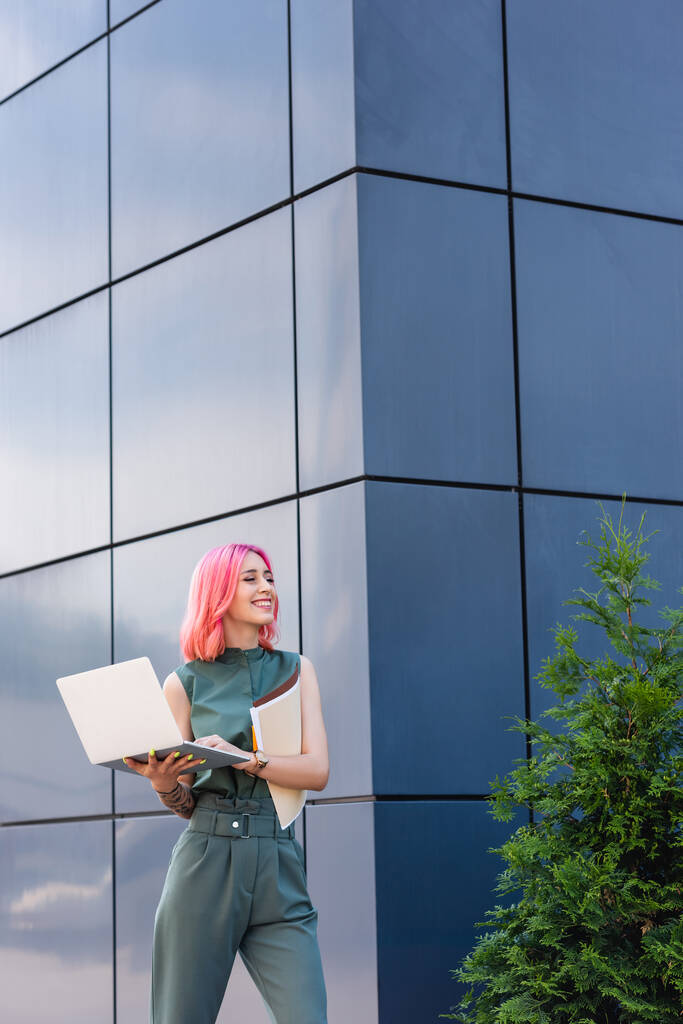 τατουάζ και χαρούμενη επιχειρηματίας με ροζ μαλλιά κρατώντας φορητό υπολογιστή και το φάκελο έξω - Φωτογραφία, εικόνα