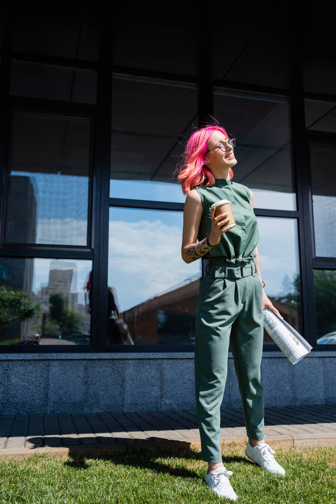 full length of cheerful businesswoman with pink hair holding rolled paper and coffee to go outside  - Photo, Image
