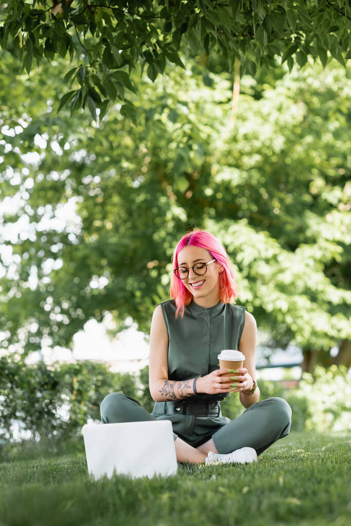 femme heureuse avec des cheveux roses et des lunettes tenant tasse en papier et regardant ordinateur portable sur l'herbe  - Photo, image