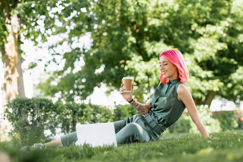 jovem alegre com cabelo rosa segurando copo de papel perto de laptop na grama  - Foto, Imagem