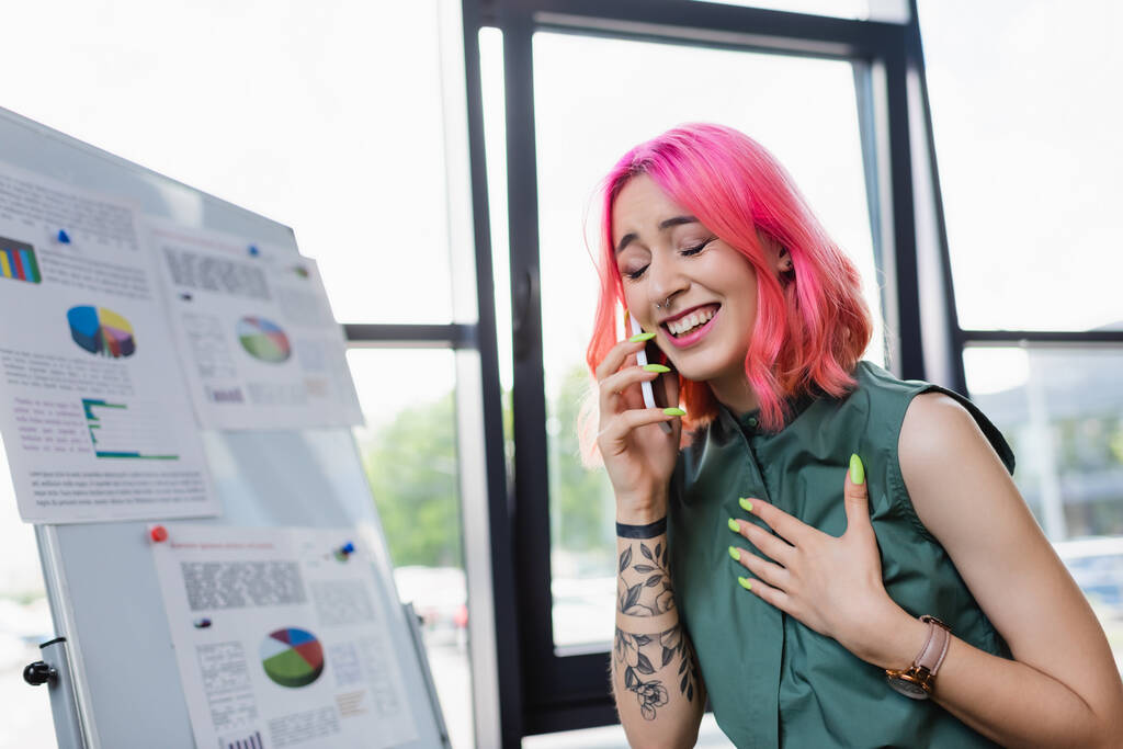 веселая и татуированная женщина с розовыми волосами разговаривает на смартфоне в офисе - Фото, изображение