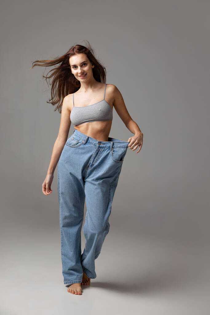 Porträt eines jungen schönen schlanken Mädchens im blauen Jeans-Overall, das isoliert vor grauem Studiohintergrund posiert. Konzept zur Gewichtsreduktion. - Foto, Bild