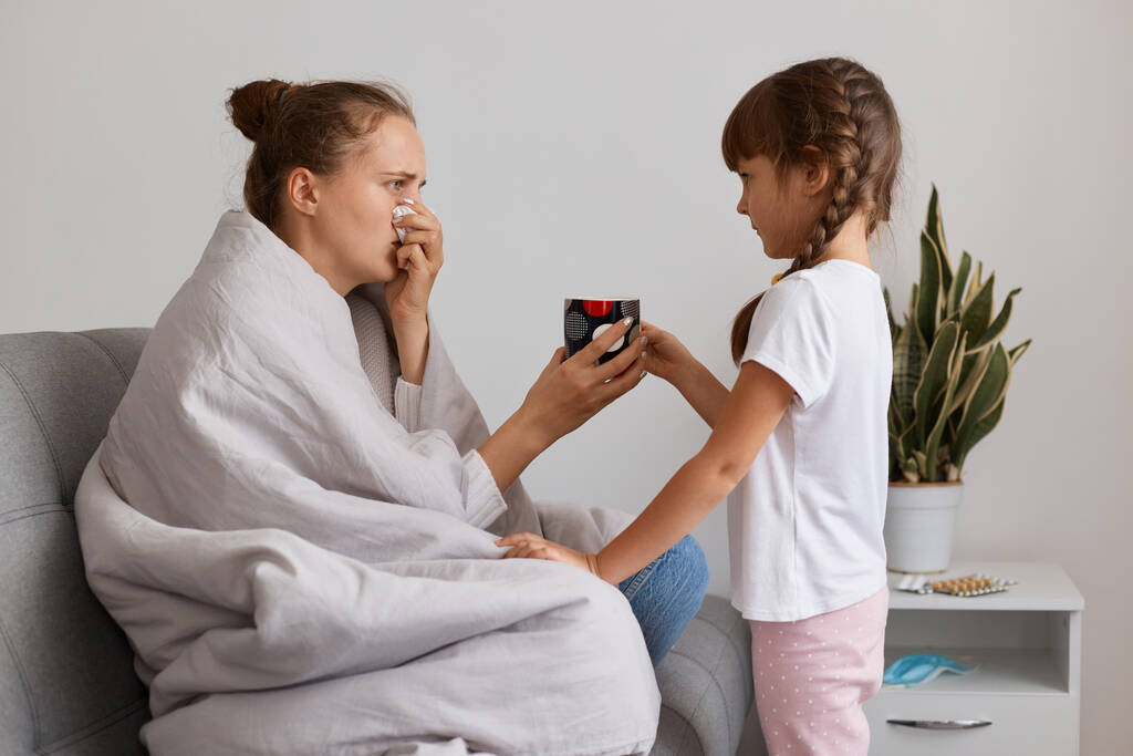 Πλάγια άποψη πορτρέτο του φροντίζοντας παιδί φέρνοντας ζεστό τσάι για την άρρωστη μητέρα της που πάσχουν από γρίπη ή πνευμονία, ποζάρουν με χαρτοπετσέτα, έχοντας καταρροή μύτη και υψηλή θερμοκρασία. - Φωτογραφία, εικόνα