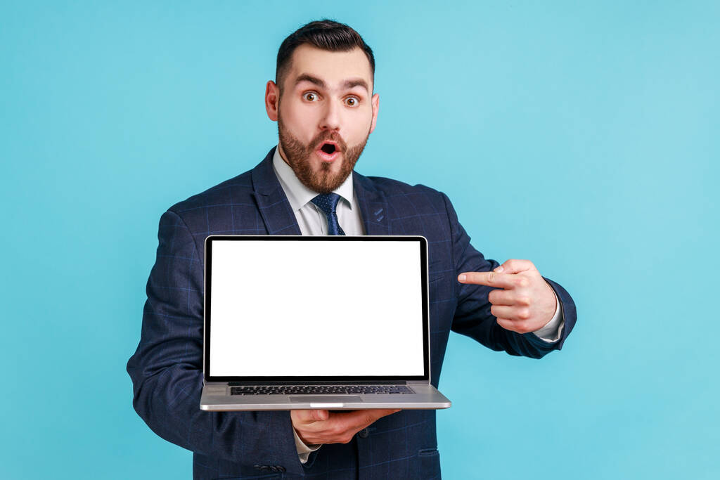 Портрет удивленного взволнованного человека с бородой в костюме, указывающего пальцем на пустой ноутбук, смотрящего большими глазами, шокированными рекламой. Крытая студия снята на голубом фоне. - Фото, изображение