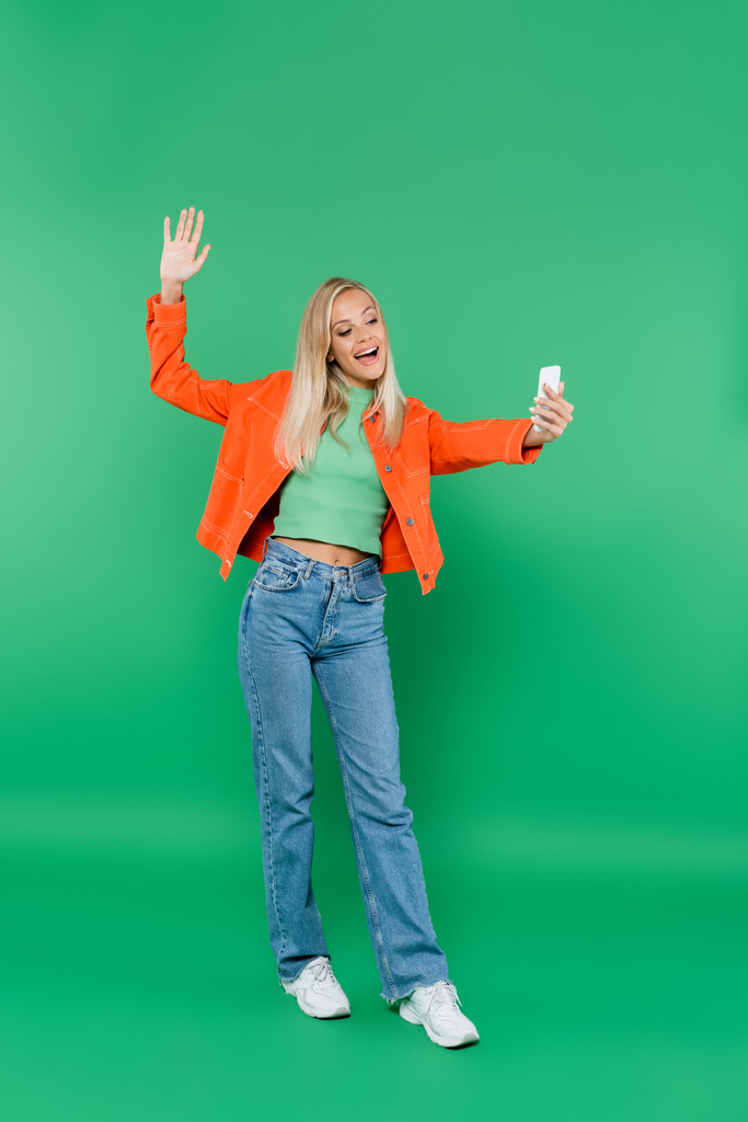 täyspitkä näkymä iloinen ja trendikäs nainen heiluttaen käsi ottaessaan selfie matkapuhelimeen vihreä - Valokuva, kuva