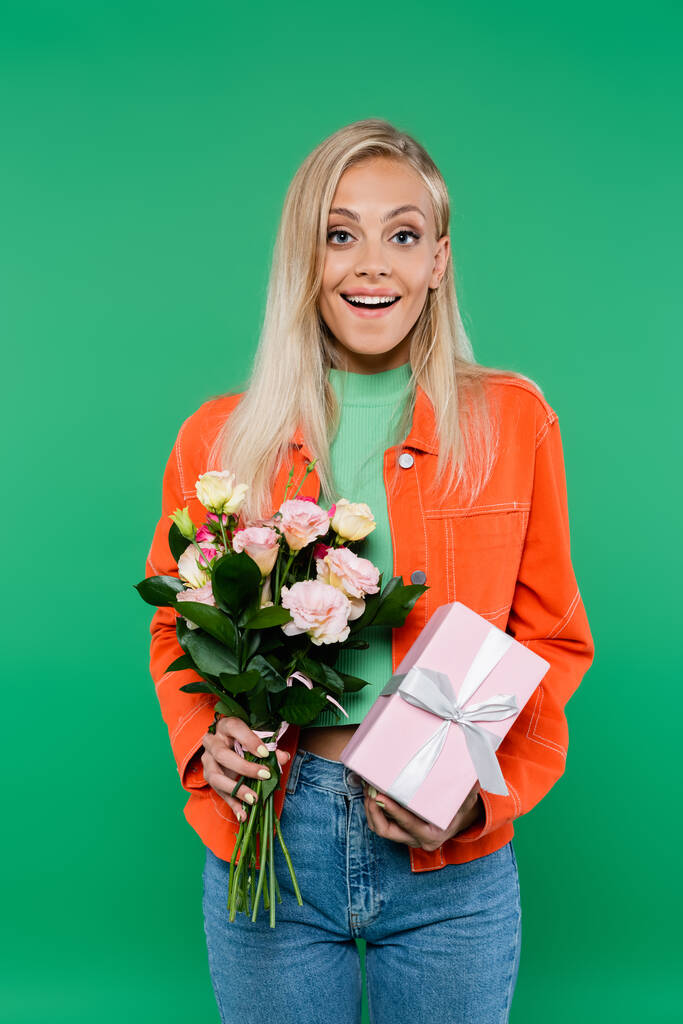 amazed blonde woman with fresh bouquet and gift box looking at camera isolated on green - Photo, Image