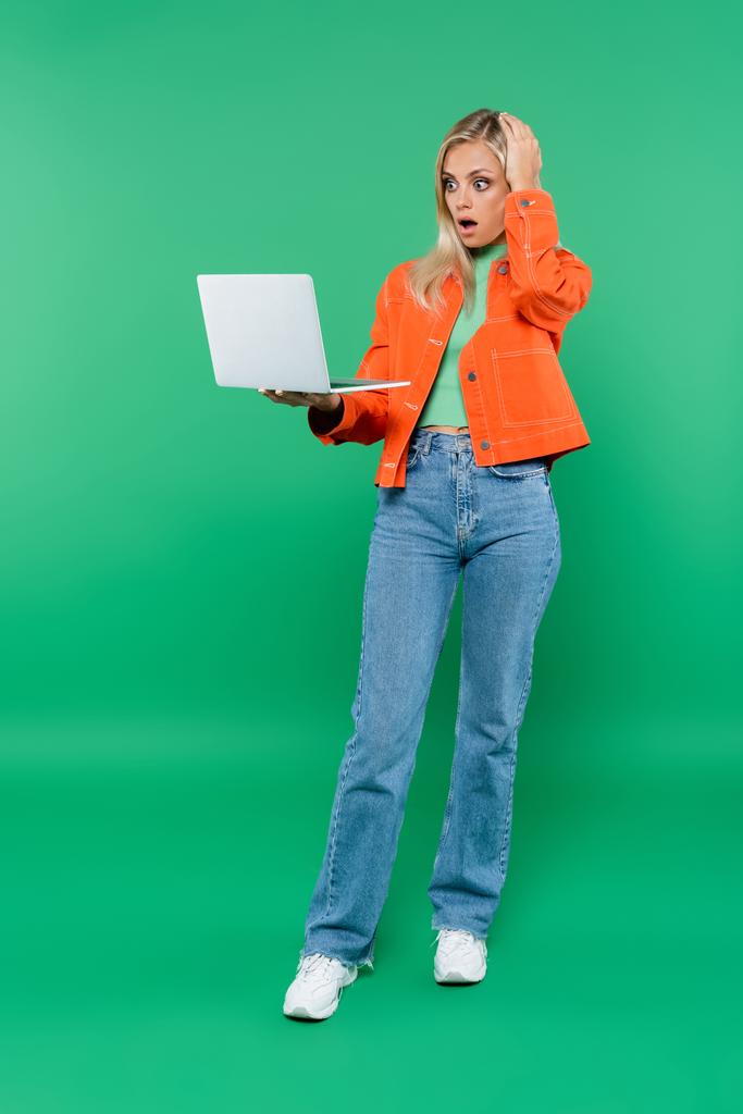 shocked woman in stylish clothes touching head while looking at laptop on green - Photo, Image