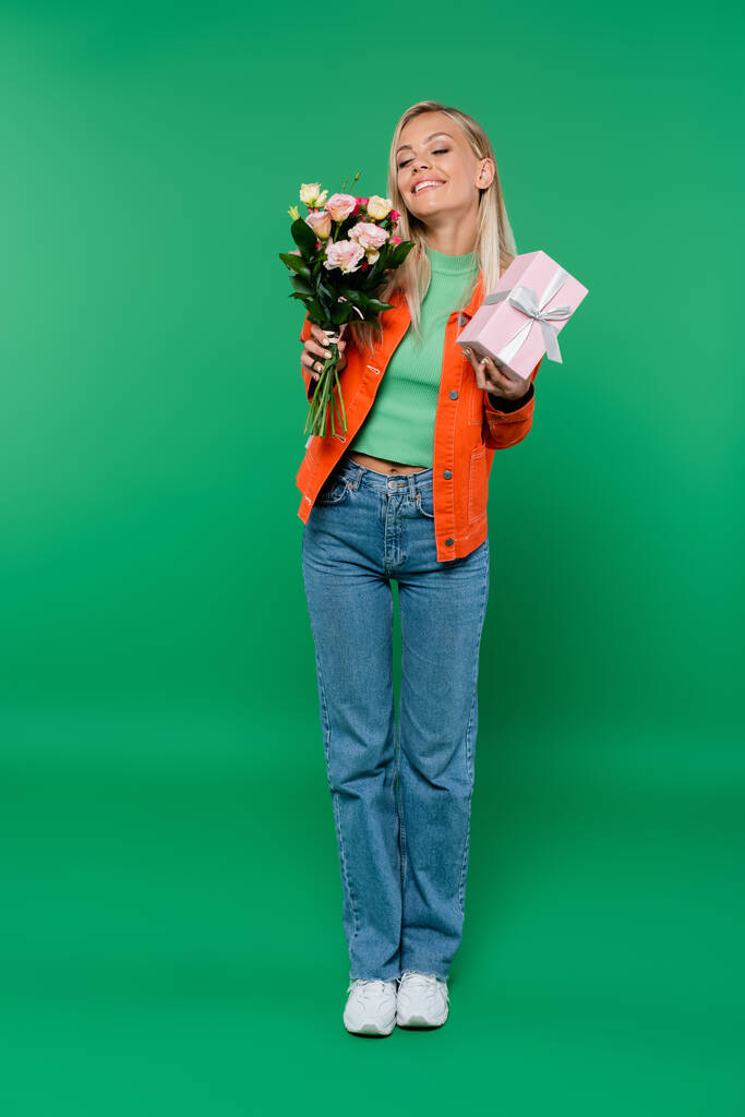 Вид в полный рост счастливой блондинки с закрытыми глазами, держащей букет и подарочную коробку на зеленом - Фото, изображение