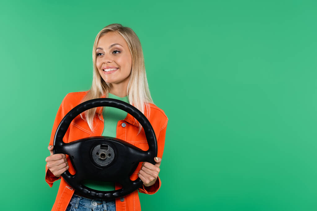 pozytywna blondynka w pomarańczowej kurtce odwraca wzrok trzymając kierownicę odizolowaną na zielono - Zdjęcie, obraz