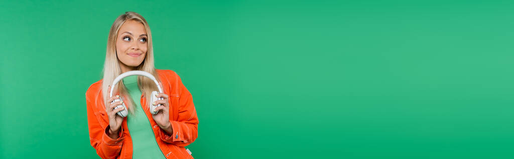 mooie blonde vrouw in trendy kleding houden koptelefoon terwijl weg te kijken geïsoleerd op groen, banner - Foto, afbeelding