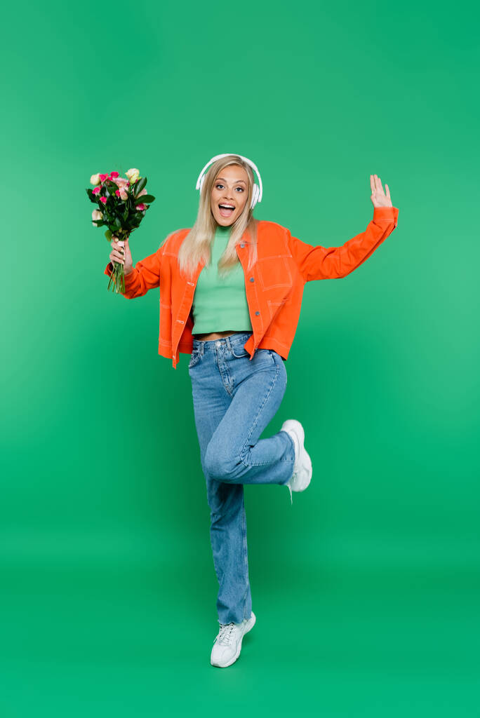 vzrušená žena ve sluchátkách a módní oblečení pózovat s květinami na zelené - Fotografie, Obrázek