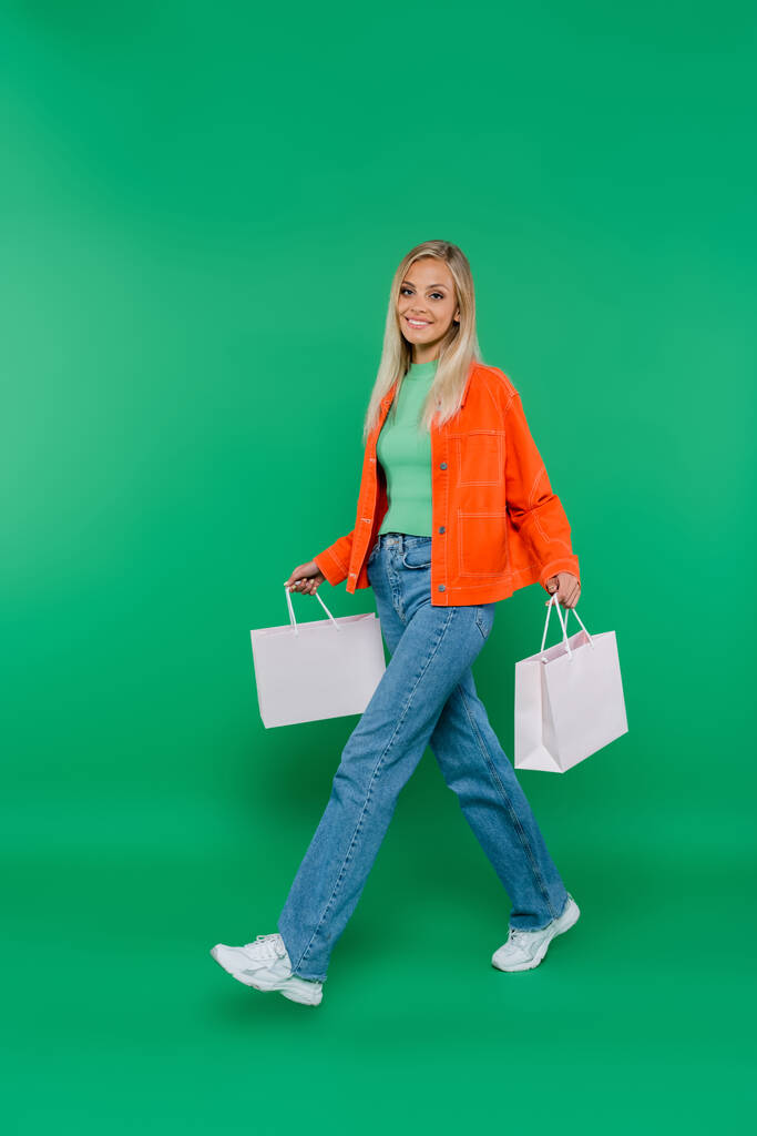 усміхнена жінка в помаранчевій куртці і джинсах, що йдуть з сумками на зеленому
 - Фото, зображення