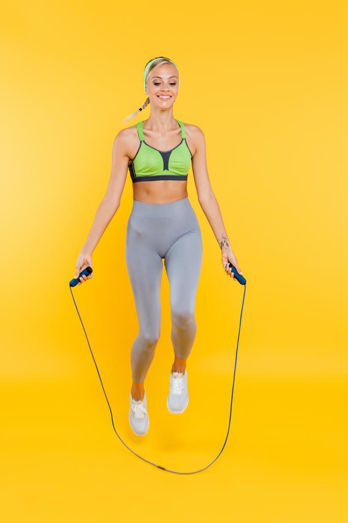 повний вигляд щасливої жінки в сірих легінсах і зелений спортивний бюстгальтер стрибає з пропусканням мотузки на жовтому
 - Фото, зображення