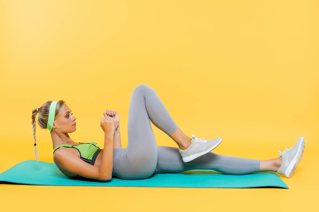 side view of woman in grey leggings and green sports bra exercising on fitness mat on yellow - Photo, Image