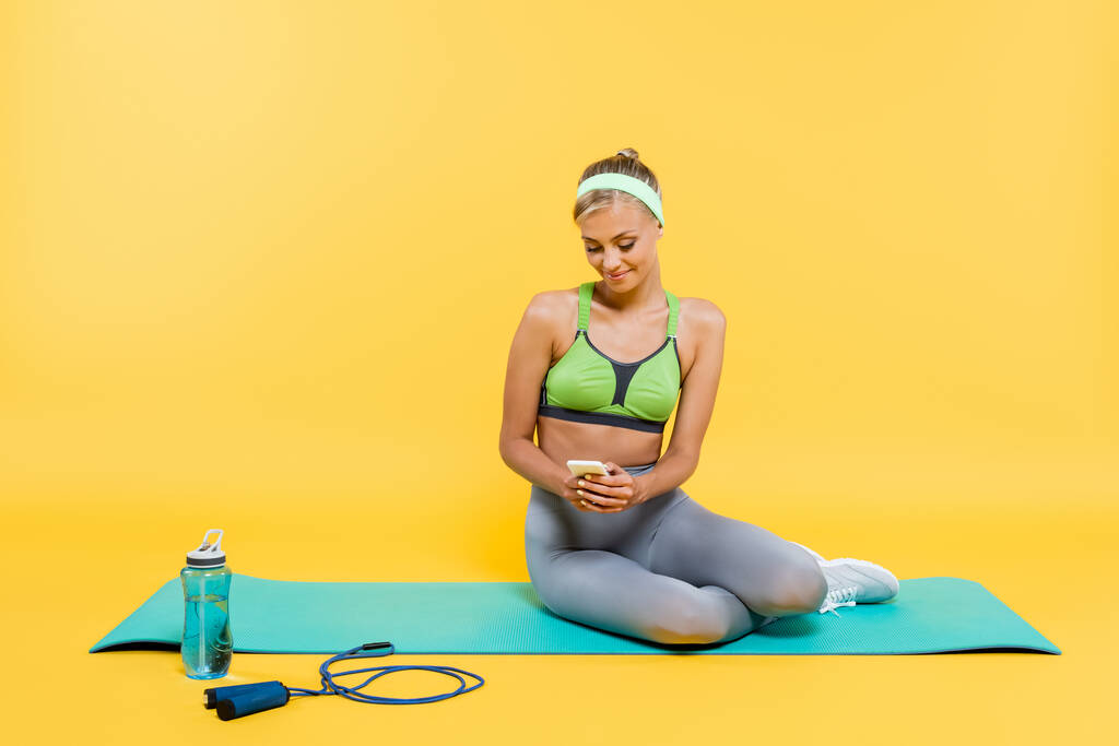 mulher satisfeita em sportswear usando smartphone enquanto sentado no tapete de fitness perto de garrafa de esportes e pular corda no amarelo - Foto, Imagem