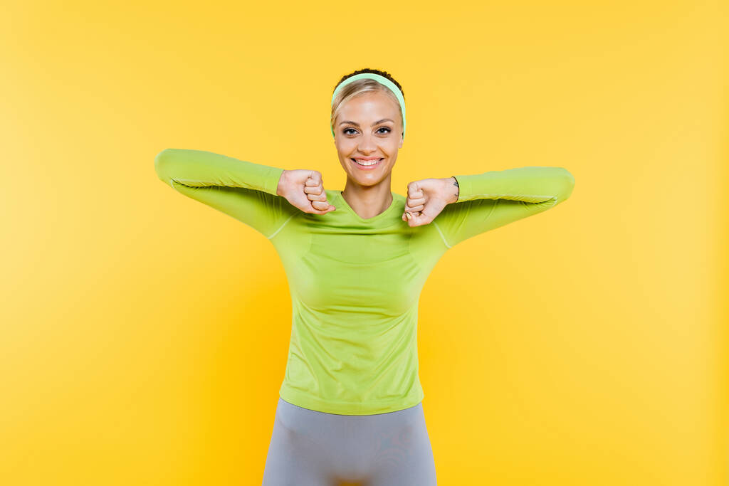 sportieve vrouw glimlachen op camera tijdens het opwarmen geïsoleerd op geel - Foto, afbeelding