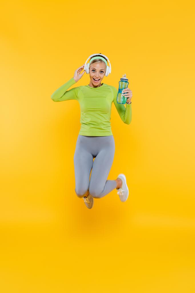 glückliche sportliche Frau mit Kopfhörer springt mit Sportflasche auf gelb - Foto, Bild