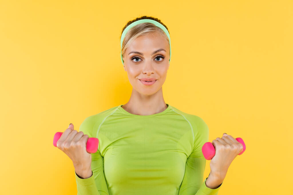 спортивная женщина упражняется с гантелями, улыбаясь в камеру изолированы на желтый - Фото, изображение