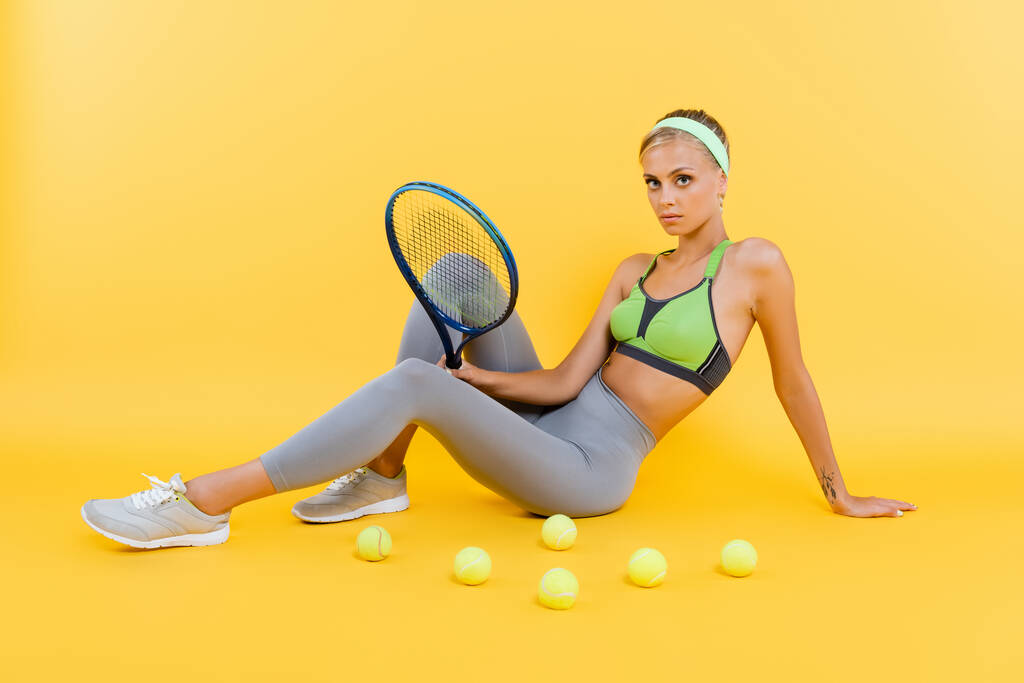 Sportliche Frau mit Schläger blickt in die Kamera, während sie neben Tennisbällen auf gelb sitzt - Foto, Bild