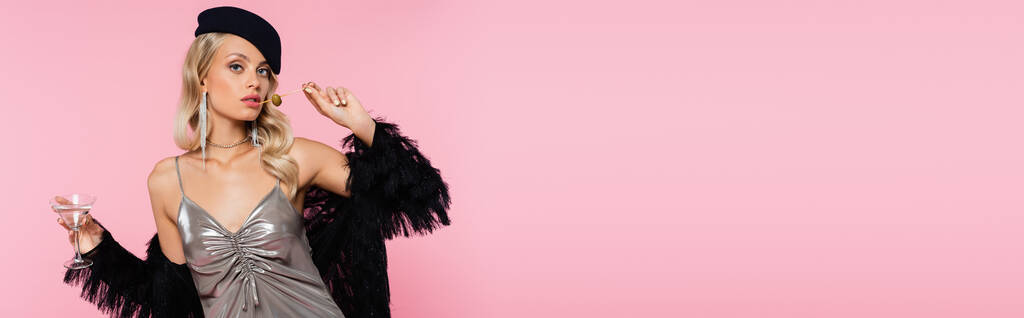 fashionable woman in black beret and faux-fur jacket holding glass and cocktail skewer with olive isolated on pink, banner - Photo, Image