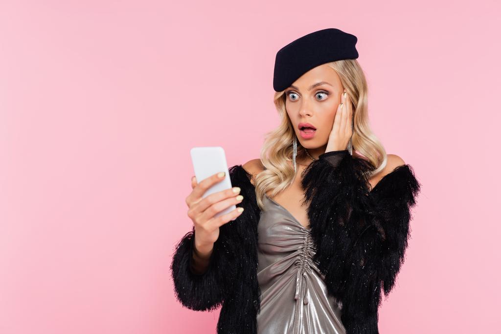 hämmästynyt nainen tyylikkäissä vaatteissa koskettaa kasvoja, kun tarkastellaan matkapuhelin eristetty vaaleanpunainen - Valokuva, kuva