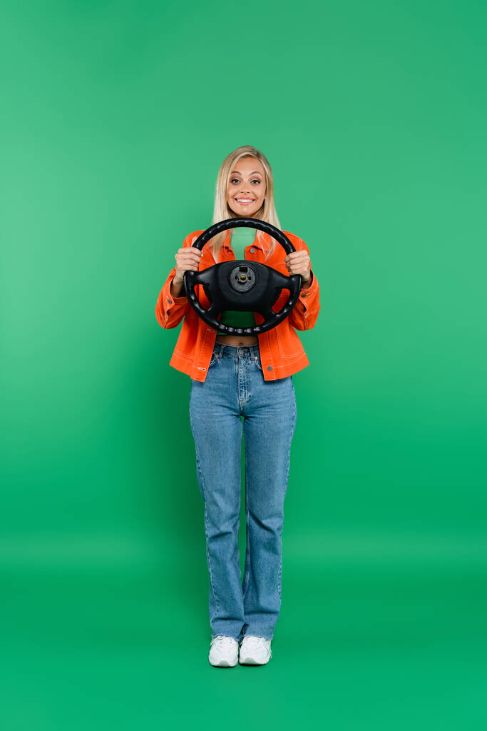 visão de comprimento total da mulher loira satisfeita em jeans posando com volante no verde - Foto, Imagem