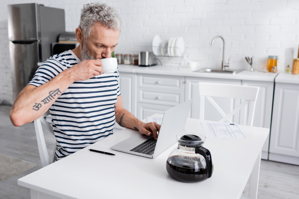 Mann trinkt Kaffee und benutzt Laptop in der Nähe von Zeitungen in der Küche. Übersetzung: "om, shanti, peace" - Foto, Bild
