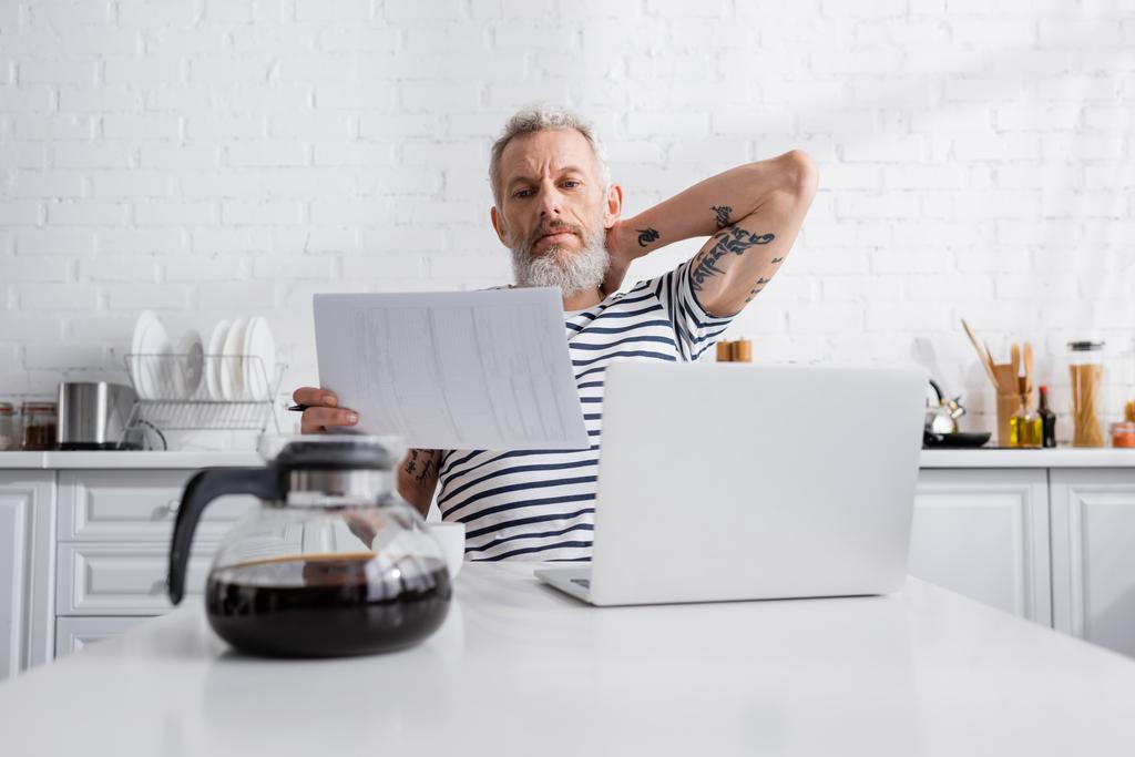 Älterer Mann mit Papieren in der Nähe von Laptop und verschwommener Kaffeekanne in Küche  - Foto, Bild