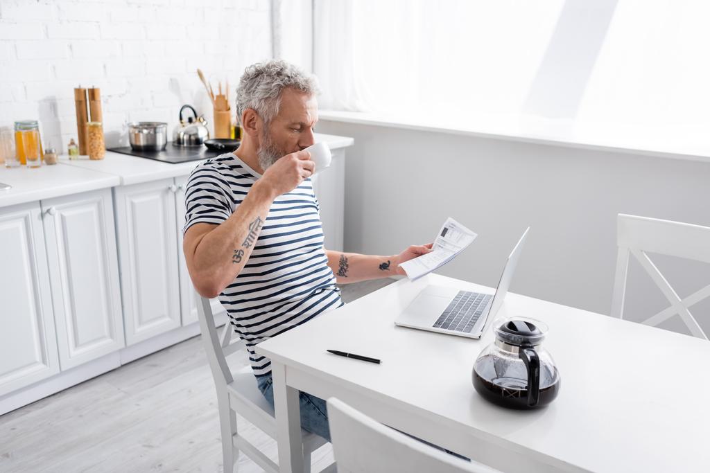 Mężczyzna trzymający papiery z rachunkami i pijący kawę przy laptopie w kuchni. Tłumaczenie: "om, shanti, pokój" - Zdjęcie, obraz