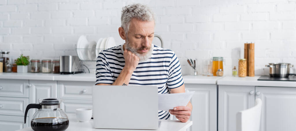 Kypsä mies raidallinen t-paita pitämällä paperia lähellä kannettavaa tietokonetta ja kahvipannu keittiössä, banneri  - Valokuva, kuva