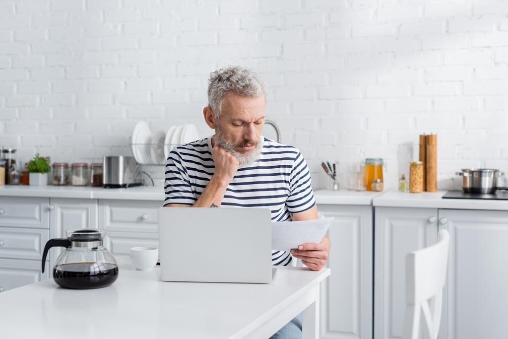 Keski-ikäinen mies etsii asiakirjaa lähellä kahvia ja kannettavaa tietokonetta keittiössä  - Valokuva, kuva