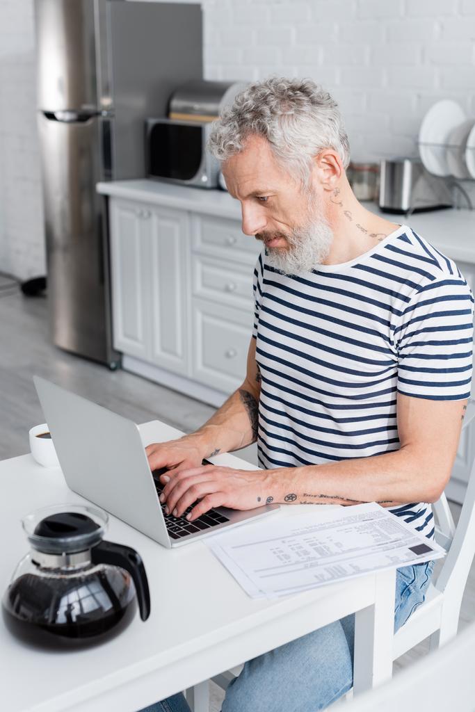 Kypsä mies käyttää kannettavaa tietokonetta lähellä asiakirjoja laskuja ja kahvia kotona  - Valokuva, kuva