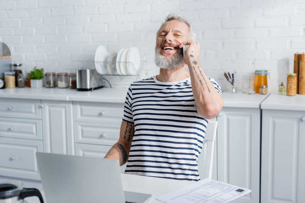 Akıllı telefondan konuşan mutlu olgun bir adam mutfaktaki faturaların yanında dizüstü bilgisayarla konuşuyor.  - Fotoğraf, Görsel