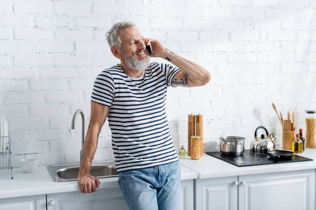 Ευτυχισμένος ώριμος άντρας μιλάει στο smartphone κοντά στον πάγκο εργασίας στην κουζίνα  - Φωτογραφία, εικόνα