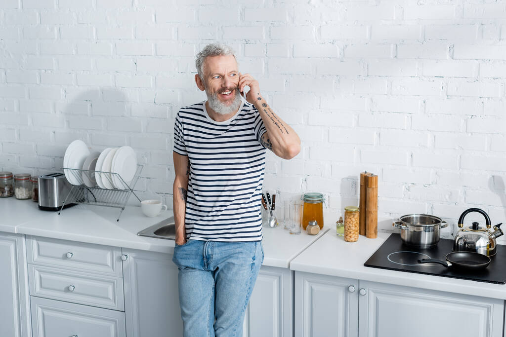 Мужчина разговаривает по смартфону возле плиты на кухне. Перевод: "Вы должны копать глубокие колодцы, если вы хотите чистой воды" - Фото, изображение
