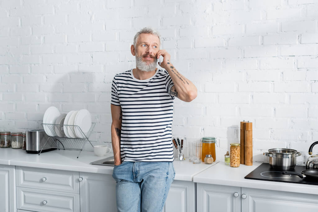 Muž středního věku v pruhovaném tričku mluví na mobilním telefonu v kuchyni  - Fotografie, Obrázek