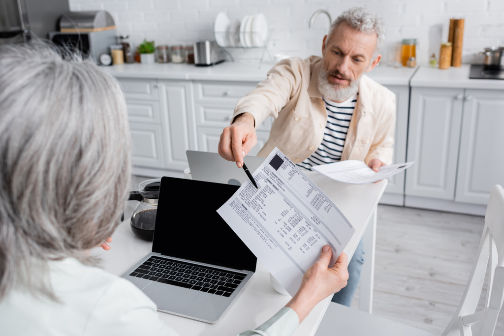 Зрелый мужчина указывает на счета рядом с женой и ноутбуки дома  - Фото, изображение