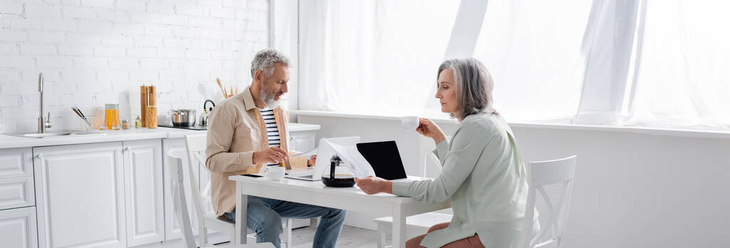 Ältere Frau hält Tasse Kaffee und Geldscheine in der Nähe Ehemann mit Laptop in der Küche, Banner  - Foto, Bild
