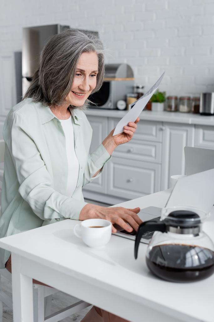 Uśmiechnięta kobieta trzyma rachunki i używa laptopa przy kawie w kuchni  - Zdjęcie, obraz